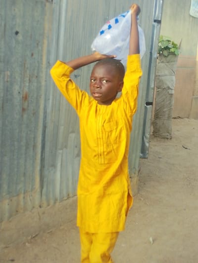 The image displays a male child standing outdoors in Babban Giji, Nigeria. The child has dark skin and short, dark hair, and is wearing a bright yellow, long-sleeved tunic with vertical dark stitching details on the chest, paired with matching yellow trousers. He stands centrally, facing forward with a neutral expression. Both hands are raised above his head, supporting a translucent plastic bag with blue markings that rests on his head. The background consists of a weathered corrugated metal surface on the left and center. To the right, a vertical structure is partially obscured by light grey, sack-like material, from which green foliage emerges at the top. The ground is unpaved, composed of light brown earth.