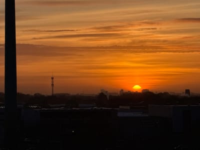 The image depicts a striking sunrise or sunset over an urban-industrial landscape, likely in Maasdijk, Netherlands, captured from an elevated vantage point. The sky is the most prominent feature, displaying a rich palette of warm colors, ranging from deep oranges and yellows near the horizon to lighter browns and grays higher up. Horizontal, streaky clouds are visible, catching the golden light.
The sun, a bright, intense yellow-orange disc, is positioned low on the horizon, partially obscured by distant landforms and atmospheric haze. Its powerful glow creates a dramatic silhouette effect for the elements below.
In the mid-ground and foreground, a dark silhouette of buildings, indicative of an industrial or commercial area, spans across the lower half of the frame. One of these buildings has the word "efficient" faintly visible on its side. A tall, slender communications antenna or cell tower is a notable vertical structure, standing on the left side of the mid-ground, silhouetted against the vibrant sky.
Beyond these structures, a line of silhouetted trees and the hazy outlines of distant city buildings are discernible on the horizon, particularly around the bright sun. The overall atmosphere is characterized by the soft, diffused light typical of dawn or dusk, creating a serene yet expansive scene.