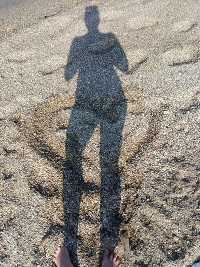 A full-body shadow of a person standing on a beach comprised of small pebbles and sand is cast on the ground. The shadow depicts the person's head, torso, arms, and legs. Visible at the bottom of the image are two bare feet, one on the left and one on the right, with toes visible. The environment suggests a sunny day. This scene is on the coast of Águilas, Spain.