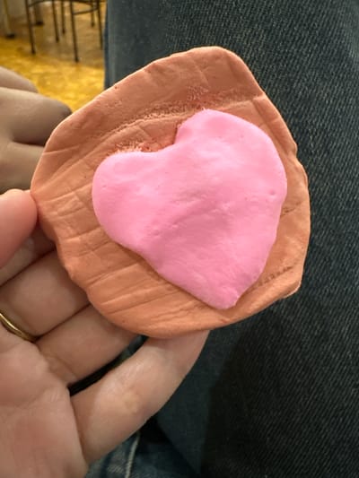 A close-up shot shows a person's hands holding a handmade object, likely crafted from modeling clay or playdough. The primary object consists of a light brown, roughly rectangular or oval base with a distinct textured, ridged surface. Layered on top of this base is a smaller, irregularly shaped blob of bright pink material, resembling an abstract form or a stylized heart.
The person's left hand, with a gold-colored ring on the ring finger, supports the object. The background primarily features the person's lap, covered in blue denim fabric, indicating a casual setting. In the lower left portion of the image, a blurred yellowish area with dark lines suggests an indoor environment, possibly a floor or furniture. The activity depicted is the holding or presentation of this craft item. No specific time of day, weather, or discernible emotions are evident from the image. There is no visible text. The scene is situated in Uva, Russia.