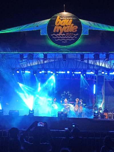 The image depicts a vibrant night scene at an outdoor stage in Kecamatan Pujut, Indonesia, during what appears to be the "Festival Pesona Bau Nyale 2025." A large, circular sign is prominently displayed above the stage, featuring the text "festival pesona" at the top, "bau nyale" in large yellow/orange letters in the center, and "2025" below it, along with wavy lines suggesting water. This sign is encircled by a bright green neon ring, matching other green accent lights on the stage structure.
The stage is brightly illuminated by numerous blue stage lights, casting strong beams through visible haze or smoke, creating a dramatic atmosphere. Overhead, metal trussing supports various lighting fixtures. On stage, four performers are visible, dressed in elaborate, possibly traditional, costumes. One central figure wears a distinctive, ornate golden headpiece. They appear to be engaged in a synchronized performance, likely a cultural or theatrical act.
In the foreground, the silhouettes of an audience can be seen, with several individuals holding up smartphones to record or photograph the event. The time of day is clearly night, characterized by the dark sky and extensive artificial lighting. The scene conveys an energetic and engaging event.