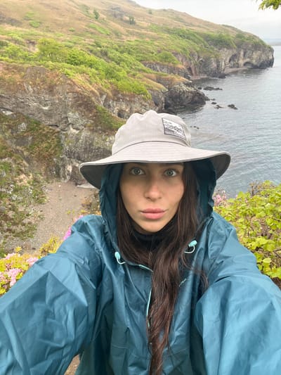 A selfie shows a woman with fair skin, green eyes, and dark hair wearing a gray hat and a dark blue rain jacket. She stands on a cliff edge, with the sea and a rocky coastline visible in the background. Green vegetation and small pink flowers grow along the cliffside. The location appears to be a coastal area, but is not identifiable as Gvozdevo, Russia.