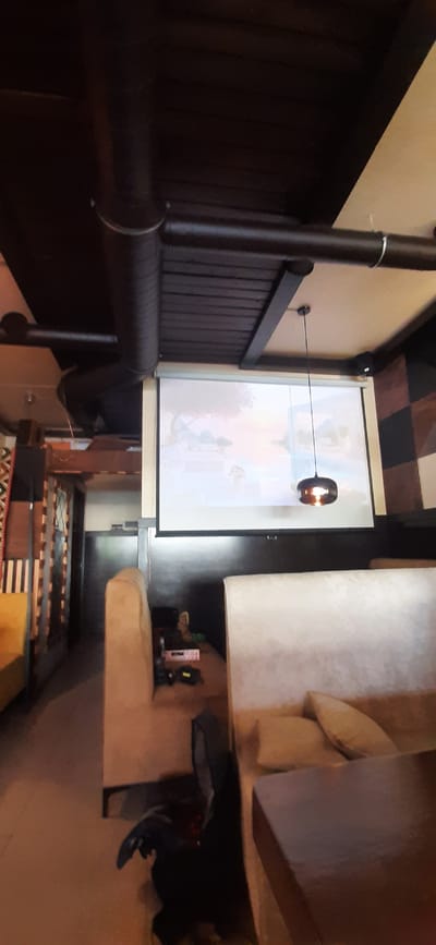 This media file shows an indoor scene, possibly a home theater or living room, with a projector screen displaying an image. The screen is large and white, with a colorful projection on it, though the content of the projection is unclear. A dark, cylindrical pipe runs vertically along the left side of the image, a common feature in industrial or older buildings. Below this pipe, there is a dark wooden wall paneling. A single light fixture hangs from the ceiling by a wire, its bulb glowing warmly. The room appears to have a sectional sofa on the right side, upholstered in a light-colored fabric. On a shelf above the sofa, several electronic devices are visible, including remote controls and what appears to be a camera or projector. The floor in the foreground shows a mat with striped patterns. The overall lighting suggests it could be evening or a dimly lit room. The setting is likely in Yessentuki, Russia, based on the provided location context. There is no visible text in the image.