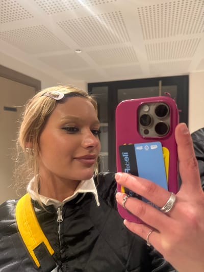 A selfie shows a blonde woman in a black jacket with yellow accents. She's holding a pink phone case with three visible camera lenses. Cards are tucked into the back of the case, including a blue debit card. The woman wears multiple rings and a barrette in her hair. The background features a white textured wall and a dark window.