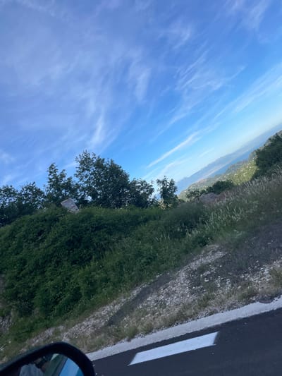 The image captures a scenic overlook near Obzovica, Montenegro, on a clear day with wispy clouds stretching across a bright blue sky. The foreground features a roadside with asphalt and a white line, bordered by a sloping embankment covered in dense green vegetation and tall grasses. In the mid-ground, a cluster of trees stands atop the embankment, partially obscuring a tilted sign with some text visible. Beyond the trees, the landscape opens up to reveal rolling hills and mountains in the distance, descending towards a body of water that appears to be the Adriatic Sea. The perspective suggests the photo was taken from a moving vehicle, possibly from the passenger seat, as indicated by the reflection in what appears to be a side-view mirror in the lower left corner of the frame. The overall scene depicts a natural, mountainous coastal environment.