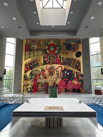 The image displays the interior of a large hall, identified as a religious space, likely a church or chapel, located within the Galil Elyon Regional Council, Israel. The focal point is a massive, multi-paneled mural covering the entire back wall, rendered in a Byzantine or Orthodox iconographic style with a dominant gold background. The mural depicts numerous stylized figures arranged in scenes and circular patterns, primarily in red, blue, green, and dark tones, illustrating various religious narratives.
In the foreground, a rectangular altar table, draped in a white cloth with black horizontal stripes along its edge, occupies the center. A small, framed religious image rests flat on the white cloth, positioned slightly to the left of the center. Two simple white candles in brass holders are visible on the altar, one near the left edge and one near the right. Directly in front of the mural, a green fabric-draped lectern or pulpit stands on the left side of the central axis. To its right, another smaller, plain wooden table is visible.
The room features a high, light gray ceiling with recessed lighting fixtures and a large central skylight, allowing natural light to enter. Tall, narrow windows on both the left and right walls extend from floor to ceiling, revealing green foliage and trees outside. The walls are constructed from light-colored, possibly stone or concrete blocks. The floor in the central area is made of polished marble, reflecting light, while the floor on the left, where rows of white metal-framed chairs are arranged, is covered in blue carpeting. Transparent plastic chairs are visible near the lectern and on the right side. No individuals are present in the scene.