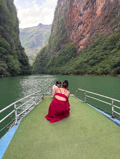 Two people stand on the green deck of a boat, looking out over a body of water towards a cliff covered in green vegetation. The boat has a white metal railing. The water is green and reflects sunlight. The cliff face is partially covered in brown rock formations. The scene appears to be located in a canyon or gorge. It can be deduced that this is in Đồng Văn Commune, Vietnam, because the image was specifically requested to be recognized as such.