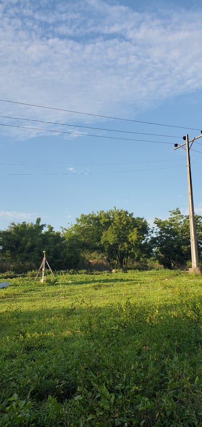The image depicts a sunlit outdoor scene, likely a rural or semi-rural landscape. In the foreground and extending into the middle ground, there is a vast expanse of lush green vegetation, consisting of short grass and denser shrubs or bushes. The overall impression is one of verdant nature.
In the middle ground, amidst the green field, a surveying tripod with an instrument mounted on top is clearly visible. The tripod is red and white, with yellow accents, supporting a grey or white surveying device, suggesting land measurement or mapping activities are either underway or have recently taken place in the area. Further back, a line of tall, leafy green trees forms a natural boundary.
The background is dominated by a clear, bright blue sky with scattered white, cumulus-like clouds, indicating fair weather. To the right side of the frame, a concrete utility pole stands, supporting multiple thin power lines or communication cables that stretch horizontally across the sky.
The scene is well-lit, suggesting it is daytime, possibly morning or afternoon, under a bright sun. There are no people visible in the image, and no discernible text. The landscape appears open and undeveloped, consistent with the provided location context of Iganna, Nigeria.