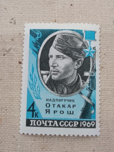 This is a photograph of a postage stamp. The stamp features a portrait of a man in a military uniform and cap, with a rifle and a medal. The text on the stamp is in Russian and includes "НАДПОРУЧИК", "ОТАКАР", "ЯРОШ", "4К", and "ПОЧТА СССР 1969". The background of the stamp is blue and white with stylized imagery of a hammer and sickle, a star, and leaves. The stamp is placed on a textured, light-colored surface. There is no visible city of Onaldy, Turkmenistan in this image.