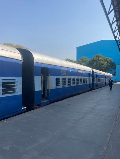 The image shows a blue and gray Indian Railways sleeper car parked on a platform in Anajpur, India. The train car has windows with bars and the word "SLEEPER" written in Hindi. A person in dark clothing is standing further down the platform near the train. A blue building and trees are visible in the background. A metal structure is overhead in the foreground. The platform is gray. The sky is blue.