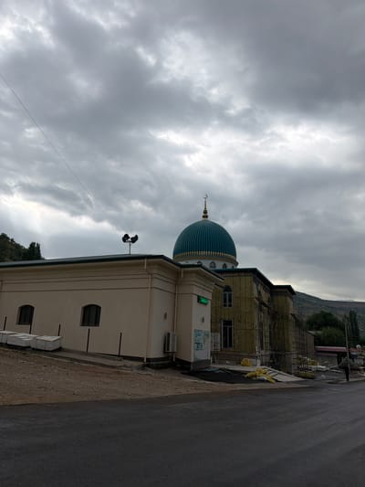 In the city of Khumsan, Uzbekistan, under a dramatic, overcast sky, a mosque is visible. The mosque features a prominent, teal-colored dome topped with a crescent moon. To the left of the dome, a cream-colored building with arched windows is situated. In front of this building, stacks of white materials are piled on the ground. Adjacent to the mosque's dome, a structure is undergoing construction, evidenced by scaffolding and insulation. A street runs across the foreground of the image, and a lone figure, carrying a large object on their back, walks away from the mosque on the right side of the frame. The surrounding environment appears hilly, with trees dotting the landscape in the background. A pair of loudspeakers are mounted on a pole atop the cream-colored building.