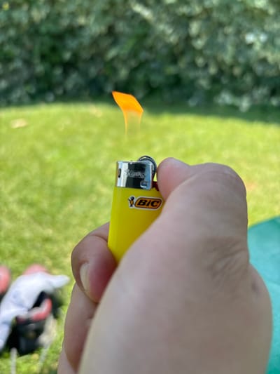 A close-up shot captures a person's hand holding a yellow Bic lighter with an active flame. The lighter is positioned to the right of the frame, with the flame extending to the left. The background is a blurred expanse of green grass and some greenery, suggesting an outdoor setting. The lighting indicates it is daytime, likely sunny due to the bright, even illumination. The scene is composed from a high-angle perspective, looking down at the hand and lighter. The focus is sharp on the lighter and the flame, while the background is intentionally out of focus. The visible text on the lighter clearly reads "BIC". The overall impression is a simple, candid moment of igniting something.