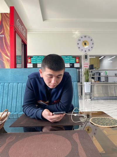 A man sits at a table, focused on a smartphone that's charging. He wears a dark blue sweater and has a name tag visible. A red Coca-Cola refrigerator sits to the left. Behind him, a wall display shows stacks of what appears to be packaged goods. A clock hangs on the wall above the display. The table has a textured brown surface and a decorative round mat. The setting seems to be a cafe or waiting area.