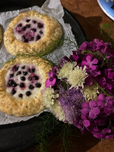 The image shows two round pastries filled with a creamy white substance and blueberries, arranged on white parchment paper. The pastries have a golden-brown crust sprinkled with poppy seeds. To the right of the pastries is a bouquet of vibrant purple and white flowers, with delicate green sprigs interspersed throughout. The scene is set on a dark surface, possibly a table. The image was taken in Velykyi Bychkiv, Ukraine, though no specific landmark or location within the city is identifiable. The focus is on the food and flowers, suggesting a still life composition.
