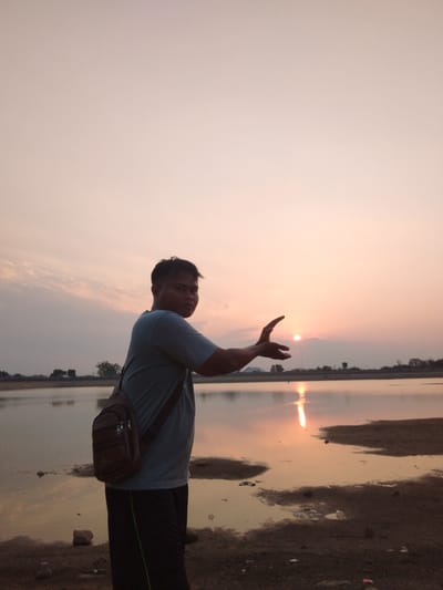 A man stands on the shore of a body of water in Sambirejo, Indonesia, facing the camera. He is wearing a light blue t-shirt and dark pants, with a brown satchel slung across his chest. His left arm is extended, with his hand cupped as if to hold the setting sun, which is partially visible on the horizon. The sky is a soft pink and orange, reflecting on the calm water. Trees and a low embankment are visible in the distance. The ground in the foreground is a mix of sand and dry earth, with some scattered debris.