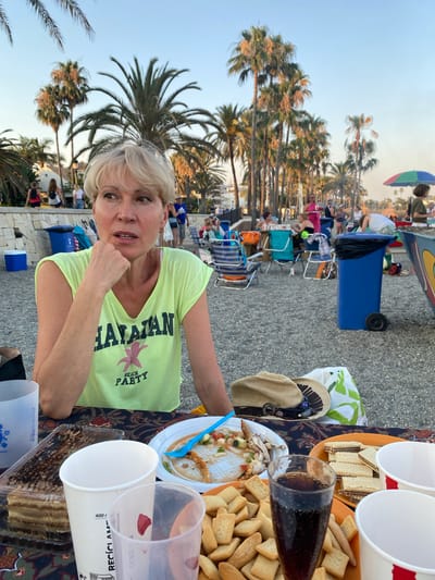 A woman with blonde hair and fair skin, wearing a yellow t-shirt with "HAWAIIAN BEACH PARTY" printed on it, is seated at a table outdoors, with a body of water and palm trees in the background. She is resting her chin on her hand, looking to her left with an engaged expression. In front of her on the table, there are various food items, including a cake, a plate of what appears to be leftovers from a meal with chicken bones, a plate of wafers, and a glass of dark liquid, likely soda. There are also several paper cups and a straw. The table is covered with a patterned cloth. Behind the table, people are visible sitting on chairs and walking, with beach umbrellas and palm trees suggesting a beachside location. The overall scene depicts an outdoor gathering or party, potentially in Marbella, Spain, given the palm trees and beach environment.