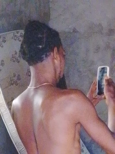 The scene is an indoor setting located in the city of Enugwu-Ukwu, Nigeria. A single individual, appearing to be an adult, is depicted from the back, shirtless. The individual has a dark skin tone and dark hair styled in short dreadlocks or twists, with a clean fade along the sides and nape of the neck. A thin, light-colored chain necklace is visible around the individual's neck. In their right hand, the individual holds a white smartphone vertically, its screen displaying an indistinct reflection. To the left, a portion of a mattress with a light-colored floral or patterned design is visible, leaning against a wall. The background consists of a textured, light grey wall, indicating an unadorned interior space with ambient, somewhat dim illumination. The individual is actively engaged with the smartphone, likely capturing an image or video, or viewing content on the device.