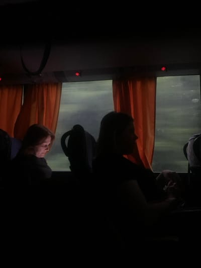 The image shows the interior of a bus. Two people are sitting in the seats, one facing the window and one facing the back of the bus. The person facing the window is a woman with dark hair, and the person facing the back of the bus is a woman with lighter hair. Both are sitting in the shadows, with only their outlines visible. The window is open and there are orange curtains on either side of the window. The window is blurry, suggesting it is raining or there is a lot of movement outside. The location of the image is in Łagów, Poland, based on the coordinates 51.382 latitude and 21.738 longitude. The image is dark, with only the outlines of the figures and the window visible. The scene is quiet and contemplative, with a sense of movement outside the bus and stillness inside. The lighting is dim, with only the faint light from the window and the overhead lights of the bus illuminating the interior. The image evokes a sense of isolation and introspection.