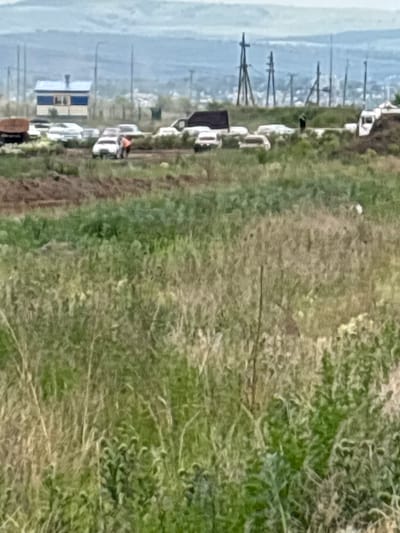 The image depicts a scene in Chernogorsk, Russia, characterized by a vast, overgrown grassy field in the foreground. In the middle ground, a cluster of vehicles is visible, including what appears to be a white SUV, several other cars, and a larger white truck with a cargo area. There are also some structures, including a small building with a blue roof and yellow siding, and several utility poles with wires in the background. The sky is overcast and hazy, suggesting an outdoor environment, possibly a construction site or an area undergoing development, given the presence of vehicles and the unfinished look of the terrain. The overall impression is one of a somewhat desolate, undeveloped area on the outskirts of a populated region.