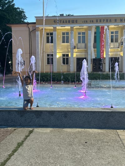 In Parvomay, Bulgaria, a young girl with dark hair is kneeling on the edge of a public fountain. She is wearing a light-colored patterned t-shirt and grey shorts, with beige sandals on her feet. Her arms are raised in the air, and she is looking towards the camera with a smile. The fountain's water is illuminated with purple and blue lights, creating a vibrant display. In the background, a yellow building with white columns stands prominently. The building has a sign with Cyrillic writing and features Bulgarian flags hanging from its facade. Trees are visible to the left of the building. The ground in front of the fountain is paved with concrete, with a line of grass growing in a crack. The scene appears to be a public square or park.