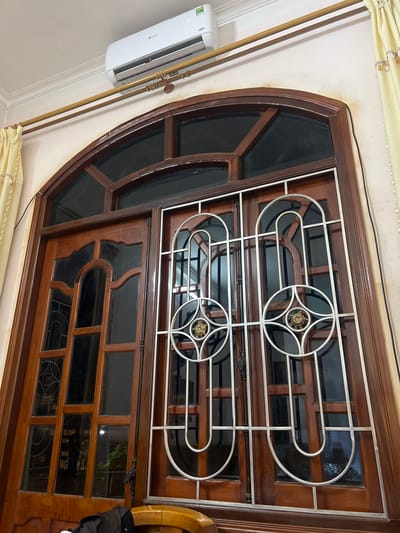 The image depicts an interior view looking upwards at an air conditioning unit mounted above a wooden arched window with decorative metal grilles. The window has multiple panes of glass, some of which reflect the exterior. Behind the window, a light-colored curtain is visible on the right side, partially drawn. The walls are painted a light color, with some yellowing or discoloration above the window frame. The air conditioning unit is white and appears to be a wall-mounted split system. The overall scene suggests a domestic or office interior in Bắc Hà, Vietnam, with architectural elements common to the region.