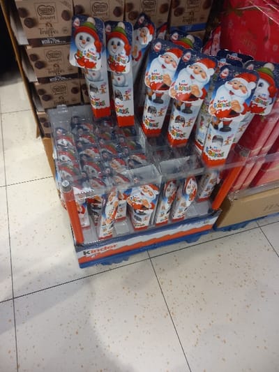 The image captures an indoor retail display of confectionery items in a store located in Sallaumines, France. In the foreground and midground, a multi-tiered point-of-sale display features numerous Kinder Surprise eggs. The upper sections of the display hold individual Kinder Surprise packages featuring festive designs, specifically smiling Santa Claus figures in red hats and snowmen wearing green and red hats, arranged upright in cardboard sleeves within clear plastic compartments. Below these, smaller, individually wrapped festive-themed Kinder Surprise items are organized in clear plastic trays on a lower shelf. The base of this display unit includes prominent "Kinder" branding in blue and red. To the left in the background, multiple brown cardboard boxes are stacked, revealing labels for "Nestlé dessert Truffes NOIR 70%" and "Nestlé dessert Lait" chocolate truffles. A portion of red packaging, likely for another festive product, is visible in the background to the right. The floor consists of light-colored speckled tiles, typical of a commercial retail environment. No individuals are present in the scene.