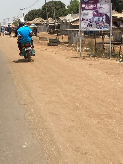 The image depicts a daytime street scene in Katcha, Nigeria. A paved road segment occupies the left portion of the foreground, bordered on the right by a wide, unpaved dirt and gravel shoulder or path. Two individuals are observed from behind, riding a green/teal motorcycle away from the viewer on the dirt surface. The driver wears a blue short-sleeved shirt, dark trousers, and a white cap. The passenger wears a dark short-sleeved shirt. The motorcycle features a visible rear tail light and license plate.

To the right of the dirt path, a large rectangular billboard stands mounted on two poles. The sign, advertising "EM & ESSENCE COSMETICS," displays images of women and various cosmetic products, listing services such as "Cosmetics," "Human Hair," "Wig Supply," "Perfumes," and "Skin Care Products." Adjacent to the billboard's base, two worn tires are stacked horizontally on the ground amidst scattered debris.

In the background, a dense arrangement of rudimentary structures with corrugated metal roofs lines the street, consistent with small shops or market stalls. Wooden utility poles carrying power lines are visible above these structures. Several indistinct figures are walking on the far left side of the paved road. The sky is bright and clear, indicating sunny weather.