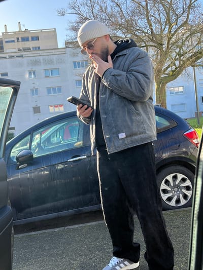 An adult male is observed standing outdoors on a paved surface under clear, sunny conditions in a residential area of Hérouville-Saint-Clair, France. He is depicted in a full-body view, oriented slightly to his right. The individual wears a light beige knit beanie, dark round sunglasses, a dark hoodie, a gray quilted jacket with buttons, black loose-fitting trousers, and light gray and white Adidas sneakers. He holds a black-cased smartphone in his left hand, directing his gaze towards its screen. Simultaneously, a lit cigarette is held between the index and middle fingers of his right hand, positioned near his mouth. To the left, the open driver's side door of a dark vehicle is partially visible. Immediately behind the man, a dark blue or black hatchback car is parked. In the background, two multi-story, light-colored residential buildings with numerous windows are visible beneath a clear blue sky. A bare deciduous tree stands to the right of the buildings.