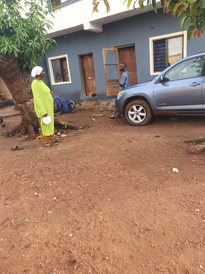 The image depicts an outdoor daytime scene in Idah, Nigeria, featuring two individuals, a car, and a building on an unpaved, reddish-brown dirt and gravel ground.
On the left, a man is seen standing next to a blue SUV, which appears to be a Toyota RAV4 with the word "LIMITED" visible above its rear wheel. He is wearing a light-colored, long-sleeved top and dark trousers. The man's face is visible, and he is looking towards the left.
To the right, a woman is captured in a lime-green long dress and a white headscarf. She is holding a small white bowl or container in her hands and facing towards the right side of the frame. Nearby, a blue plastic chair is present, bearing the word "TARA" on its backrest.
A large tree with green leaves, possibly a mango tree, stands prominently in the foreground on the left. In the background, a building with dark grey walls is visible, featuring several wooden-framed windows and wooden doors. The bright lighting suggests it is daytime with fair weather.
