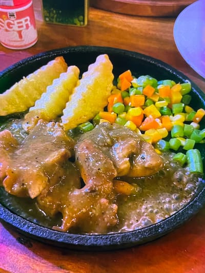 This is a close-up, overhead shot of a sizzling steak dish served on a black cast-iron platter, placed on a wooden surface. The platter contains pieces of steak in a rich, brown gravy, accompanied by a medley of diced vegetables, including green beans, carrots, and yellow bell peppers. Three golden-brown potato wedges with ruffled edges are arranged on the side.

In the bottom left corner, a partially visible red and white cylindrical object, possibly a can or a small container, bears some text in what appears to be an Asian script, with the characters "F" and "T" discernable. To the left of this object, a small green rectangular item, possibly a phone or a coaster, is also visible.

The overall scene suggests a meal being served, likely at a restaurant given the presentation on a sizzling platter and the presence of accompanying items. The lighting suggests an indoor setting, possibly with artificial light, as there are no clear indications of time of day or weather. The image is primarily focused on the food, highlighting its texture and colors. The focus is sharp on the food, with a slight blur in the background, emphasizing the culinary subject. No people are visible in the frame. The cuisine appears to be a Western-influenced dish, possibly adapted to Indonesian tastes, given the provided location context of Cadek, Indonesia.