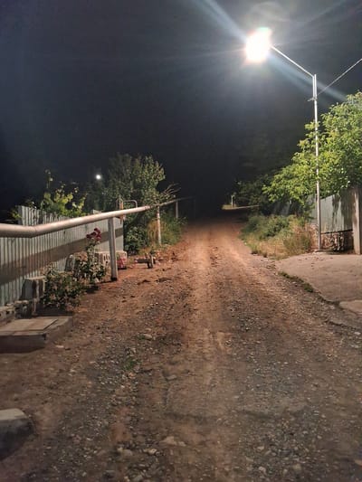 The image depicts a dirt road at night in Yeghegnadzor, Armenia, illuminated by a streetlamp. The road is unpaved and appears to be uneven, with scattered gravel and stones. On the left side of the road, there is a light-colored metal fence and some vegetation, including rose bushes with red and pink flowers. Several concrete blocks are visible near the fence. On the right side, there is a light-colored metal fence with vertical slats, and green trees and bushes. The streetlamp is positioned on the right side of the road, casting a bright light and creating a lens flare effect. The road recedes into the darkness in the distance, where another smaller light is visible. The overall impression is of a rural or suburban setting at night.