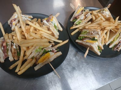 Two black plates, each containing two halves of a club sandwich and a generous portion of french fries, are positioned on a scratched metallic surface. The sandwiches appear to be made with toasted bread, lettuce, tomato, and a fried egg. Several wooden toothpicks are used to hold the sandwich halves together. The plates are slightly offset, with the left plate closer to the foreground. The overall setting suggests a food establishment, possibly a restaurant or cafe, located in Sotira, Cyprus. There are no people visible in the image.