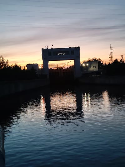 This media file depicts a concrete structure, likely a lock or dam, on a body of water at dusk. The sky displays a gradient of colors, transitioning from a pale blue at the top to warm orange and yellow hues near the horizon, indicating either sunrise or sunset. Several horizontal power lines are visible stretching across the sky.

The central subject is the imposing concrete structure, which has an open gateway. Lights are visible within and around the structure, casting reflections on the water's surface. To the right of the structure, there is a smaller, light-colored building with illuminated windows. Behind the main structure and the building, a dense line of trees forms a dark silhouette against the colorful sky. A tall, lattice-style power pylon is also visible on the right side of the frame.

The water in the foreground is dark and rippled, reflecting the colors of the sky and the lights from the structure, creating elongated, shimmering patterns. The overall mood is serene and quiet, characteristic of the transition between day and night. The scene suggests an industrial or infrastructural setting, likely a waterway used for navigation or water management. No people are visible in the media file. The time of day is clearly dusk, with the atmospheric lighting being a prominent feature. There is no visible text in the media file.