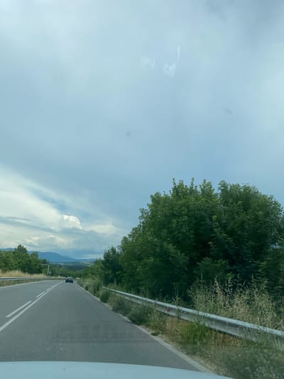 The image shows a view from a vehicle driving on a road. The road is asphalt, with double white lines indicating two lanes. To the right of the road is a metal guardrail, bordered by dry grass and green foliage. A large, dense green tree is prominent on the right side of the frame. In the distance, mountains are visible under a cloudy sky with some patches of blue and a large, white cloud formation. A dark-colored car is traveling in the same direction on the road ahead. The scene appears to be in a rural or semi-rural area.