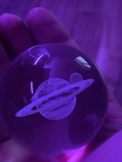 The image shows a person's hand holding a clear glass sphere with an engraved image of Saturn and its rings. The sphere is illuminated by purple light, which casts a purple hue on the hand and the background. The engraving of Saturn is visible inside the sphere, with its distinct rings and a smaller moon. The lighting creates a dramatic and somewhat mystical atmosphere. The scene appears to be indoors, given the close-up focus on the hand and the object, and the absence of any outdoor environmental cues. The time of day and weather are not discernible. There is no visible text in the image.