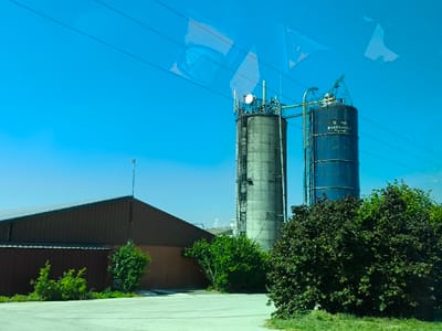Two industrial silos stand tall against a clear blue sky. The silo on the left is light gray and equipped with antennas and a ladder. The right silo is dark blue, labeled with white text, and connected to the other by a pipe. In the foreground, a low-slung brown building sits adjacent to green shrubbery. Overhead, power lines stretch across the scene. Reflections are visible in the glass through which the photograph was taken.