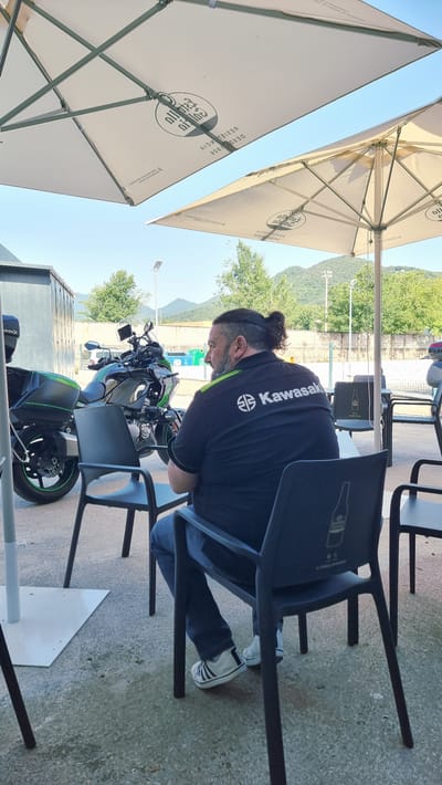 A man with his hair pulled back in a bun is sitting in a black plastic chair at an outdoor cafe in Olot, Spain. He is wearing a black polo shirt with a green stripe and "Kawasaki" logo, dark blue jeans, and white sneakers. He is facing away from the camera, looking to his left. Next to him is a black and green Kawasaki motorcycle. Two large white umbrellas are providing shade over the cafe seating area. The cafe is located on a concrete patio, with a fence and trees in the background. The sky is clear and blue.
