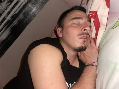 A man is asleep on a bed. The man has dark hair, a beard, and is wearing a black t-shirt. He is lying on a pillow with a red and white patterned pillowcase. His left arm is bent and his hand is near his face. The background includes a wall and a piece of furniture with a black and gray pattern. The scene does not seem to be in the city of Tulovo, Bulgaria.