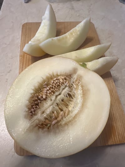 A half of a honeydew melon and several wedges of the same melon are arranged on a wooden cutting board. The half melon is on the lower left of the board and shows the white flesh and the seed cavity filled with seeds. The wedges are stacked on the right side of the board, with one on top and two below. The cutting board is on a countertop with a marbled pattern. The image was taken in Весна-53 Снт, Russia.