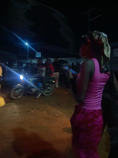 In the city of Sauka, Nigeria, at night, a young woman stands facing away from the camera, wearing a pink tank top and a patterned skirt. Her hair is wrapped in a patterned scarf. She holds a smartphone in her right hand, and her left arm is bent at the elbow. In the background, a motorcycle is parked, and other individuals are visible in the dimly lit street. A streetlamp illuminates the scene with a bright blue light. The ground appears to be unpaved.