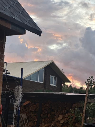 The image depicts a rural scene in the village of Tumilavichy, within the Tumilavichy rural council of Belarus. In the foreground, a substantial pile of split firewood is stored under a dark, open-sided shelter. Above the firewood, the dark wooden facade of a house is visible, featuring a white-framed window. To the left, part of another brick building is seen, with a string of decorative lights hanging from a blue pole. The middle ground is dominated by the roof of a dark wooden house with a light-colored, corrugated metal roof, and a prominent gable with windows. The background is filled with a dramatic sky at dusk, characterized by soft pinks, oranges, and grays in the clouds, suggesting the aftermath of a sunset. The overall atmosphere is quiet and tranquil, typical of a rural setting.