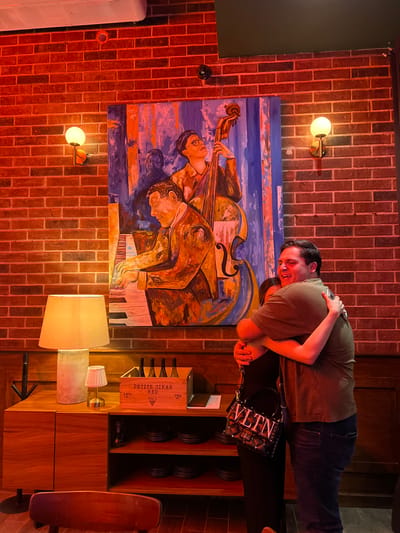 An indoor scene features a person embracing another, standing in front of a colorful, music-themed painting against a red brick wall. The visible person is a young adult with dark hair, wearing a dark olive t-shirt and blue jeans, smiling broadly while hugging someone whose arm is visible.

The large painting, rendered in a cubist or expressionistic style with blues, oranges, and browns, depicts three figures playing musical instruments: a cello or double bass, and a piano.

The setting is warmly lit by two wall-mounted globe lights and two table lamps. Below the painting, a wooden cabinet displays a wooden wine box labeled "PETITE SIRAH RED 1963 12% VOL. 750 ML" along with wine bottles, and several dark, circular objects. A black handbag with the prominent text "VLTN" and studded details rests on the cabinet. A wooden skateboard leans against the cabinet on the right.

A white air conditioning unit is visible on the left side of the brick wall, and a small black security camera is mounted high on the wall. The warm artificial lighting suggests an evening or nighttime setting. The overall ambiance is cozy and artistic, possibly in a restaurant, bar, or home entertainment area in Mérida, Mexico.