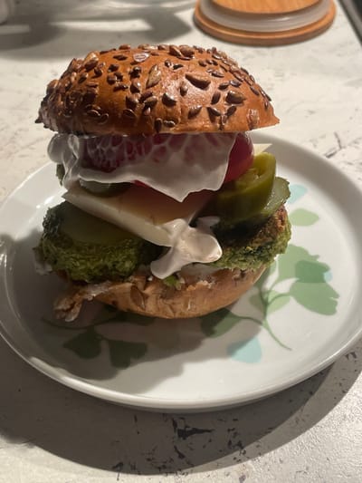 A vegetarian burger is on a white plate with a leafy green design. The burger consists of a round bun covered in sesame seeds, two green patties, a slice of cheese, a slice of tomato, a pickle, and a dollop of mayonnaise. The plate is resting on a light grey surface. The coordinates 55.659, 37.591, place the scene in the city of Moscow, Russia.