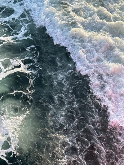 An overhead view depicts turbulent ocean water in Kato Karioutes, Greece. The scene is dominated by a dynamic interplay of deep blue-green water and extensive white foam from breaking waves. A prominent diagonal line of white, frothy foam extends from the upper right to the lower left, marking the crest of an agitated wave. This foam is brightly illuminated by sunlight, exhibiting stark white highlights and subtle pinkish-purple undertones, particularly on the right side of the frame. To the left of this main wave, the water is darker, displaying intricate patterns of smaller foam patches and disturbed surface ripples. No individuals, vessels, or landforms are present within the frame. The environment is exclusively marine, focusing on the immediate water surface.