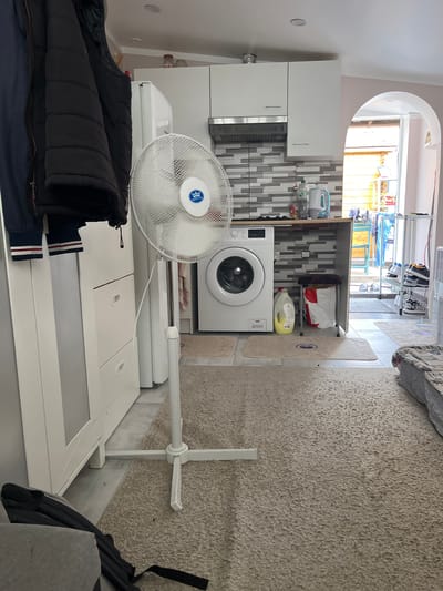 The image depicts an indoor scene, likely a laundry area or kitchen adjacent to a living space, located in the city of Stains, France. A white standing fan is positioned in the foreground, facing the viewer. To its left, a tall, white cabinet or appliance, possibly a refrigerator or wardrobe, is visible, with two dark jackets hanging on its side. Behind the fan, a white front-loading washing machine is present. The background reveals a kitchen area with white cabinets, a tiled backsplash, and a countertop. Beyond the kitchen, an open doorway leads to an outdoor space with wooden structures and laundry on display. The floor is covered with a beige carpet in the foreground and tiled in the background. No people are visible in the scene.