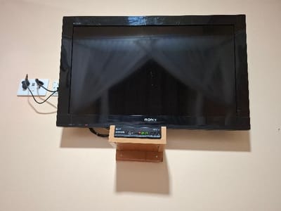 The image shows a black flat screen television mounted on a light beige wall in a residential dwelling in the Karagita neighborhood of Nairobi, Kenya. The television is a Sony brand and is mounted on a wooden shelf.  A black cable is connected to the television and leads to a white electrical outlet on the wall, where it joins another black cable. The television screen is black and off, showing a reflection of the white ceiling above.  The digital clock on the attached black cable box reads 15:21.  The room appears to be a bedroom.