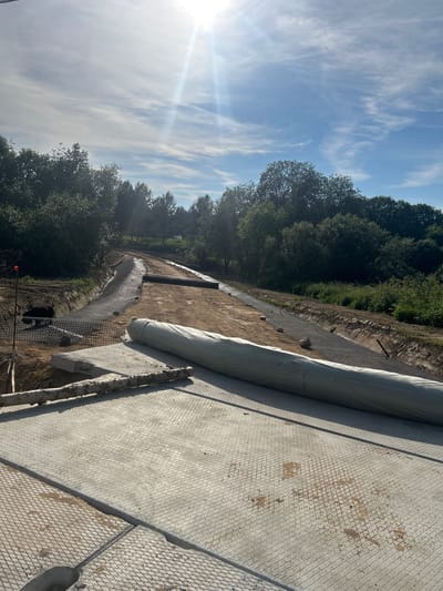 This image depicts a construction site, likely in Svetloe, Russia, with a focus on erosion control or slope stabilization. The scene is outdoors, under a partly cloudy sky with bright sunlight. In the foreground, several large, diamond-patterned concrete slabs are laid out, suggesting a temporary pathway or work platform. A large, rolled-up grey tarp or geotextile material lies on these slabs, positioned along the right side of the frame.
The middle ground reveals a sloped area being prepared for construction. A mesh or geotextile fabric, possibly for soil reinforcement or drainage, is spread across the slope and down into a trench-like excavation. The soil appears to be a mix of sand and gravel. Several large stones are visible on the fabric. Along the left side of the slope, a black mesh netting is installed, possibly to prevent debris from falling. A red marker is planted in the ground near the netting.
The background is dominated by a dense line of lush green trees, forming a natural backdrop to the construction activity. The overall impression is one of ongoing earthwork and infrastructure development in a natural environment. There are no people visible in the image.