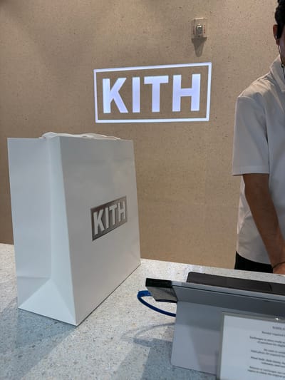 In Miami Beach, United States of America, a retail store's checkout counter is depicted. The counter is made of speckled terrazzo material. To the left, a white shopping bag with the "KITH" logo prominently displayed in a silver rectangular box is visible. Behind the bag, a wall with a speckled texture features a lit "KITH" sign. A fire alarm is mounted on the wall above the sign. On the right side of the counter, a person wearing a white polo shirt and black pants stands, partially obscured from view. A tablet device and a receipt are present on the counter.