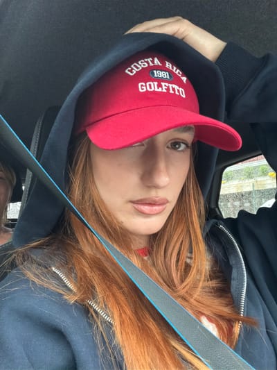 A woman with long red hair wearing a red baseball cap with white lettering that reads "Costa Rica 1981 Golfito" is wearing a dark blue hoodie. A blue seatbelt runs diagonally across her body. The setting appears to be inside a car. Outside the car window, it appears to be raining in an urban setting, suggesting a possible location in Spreitenbach, Switzerland.