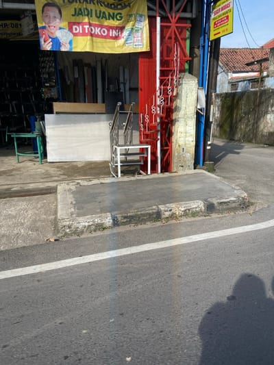 A street scene in Purwakarta, Indonesia (latitude: -6.56, longitude: 107.437) shows a sidewalk with a concrete curb and a set of metal stairs leading up to a red-painted steel structure attached to a building. The structure appears to be a fire escape. A large white sheet of material leans against the building wall. A sign with the words "JADI UANG DI TOKO INI" hangs above the entrance. The street is paved with asphalt and a white painted lane line. A shadow of a person is cast on the street. There are no people visible in the image. The scene is illuminated by bright sunlight.